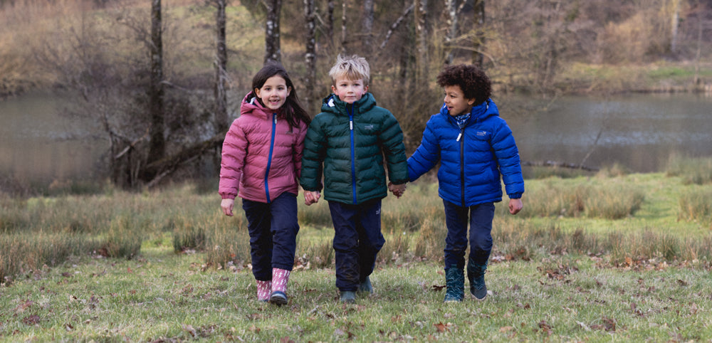 Three children in colorful waterproof jackets and rain boots walking outdoors by a lake