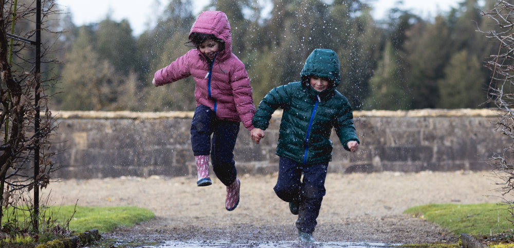boy and girl holding hands and jumping in the rain wearing colourful muddy puddles puffer jackets and wellies