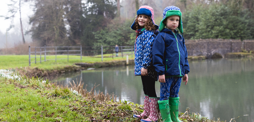 boy and girl stood by lake wearing muddy puddles waterproof jackets, wellies and trapper hats