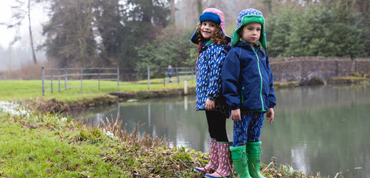 boy and girl stood by lake wearing muddy puddles waterproof jackets, wellies and trapper hats
