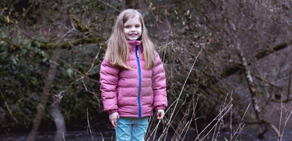 girl stood in woodland setting wearing muddy puddles pink puffer jacket and thermals