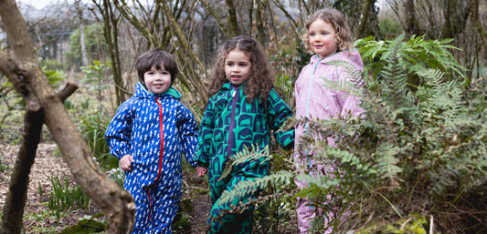 Three children in colorful waterproof rain suits playing outdoors in a wooded area