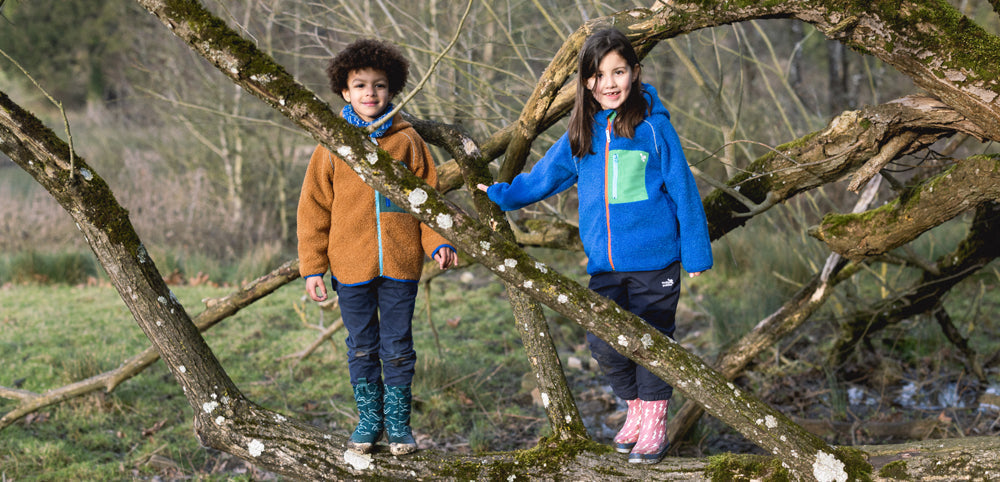boy and girl standing on tree branch wearing muddy puddles borg fleece jackets and wellies