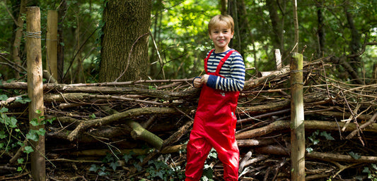 boy stood by handmade woodland fence, wearing red muddy puddles waterproof dungarees