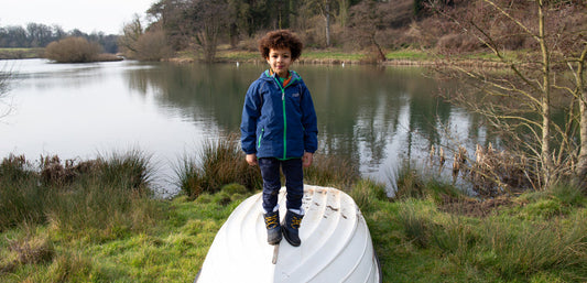 Child in waterproof outdoor jacket and boots standing on upturned boat by a lake