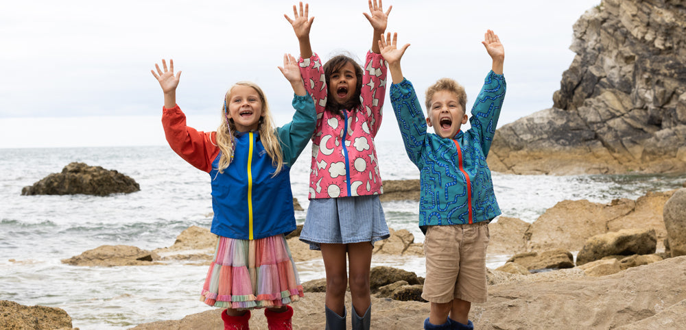 children at beach with arms in the air wearing muddy puddles waterproof jackets