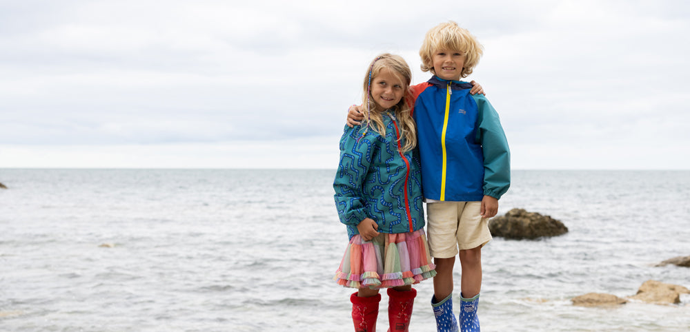Two children in colorful waterproof jackets and rain boots standing by the sea
