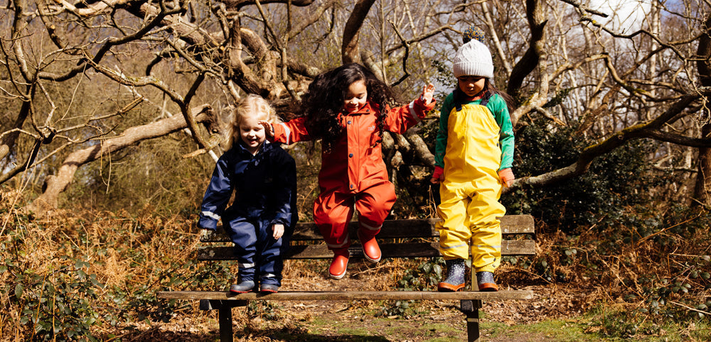 children jumping off bench in woodland area, wearing colourful muddy puddles waterproofs