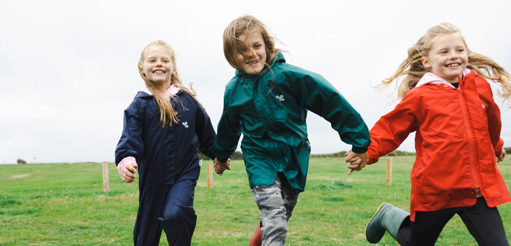 children running through field holding hands, wearing colourful muddy puddles waterproof jackets