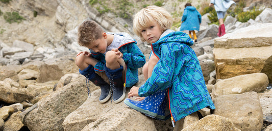 two boys sat on rocky beach wearing bold muddy puddles raincoats and wellies