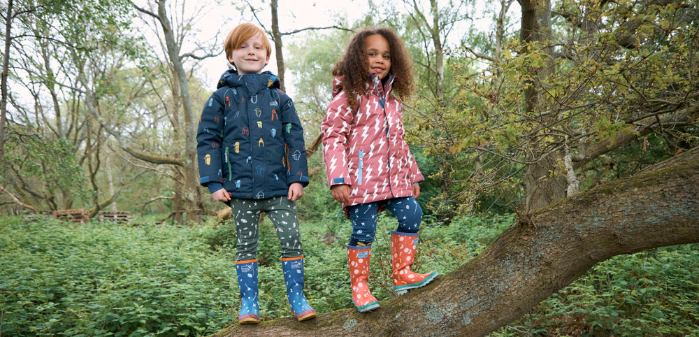 children stood on tree branch wearing vibrant muddy puddles ski jackets, thermals and wellies