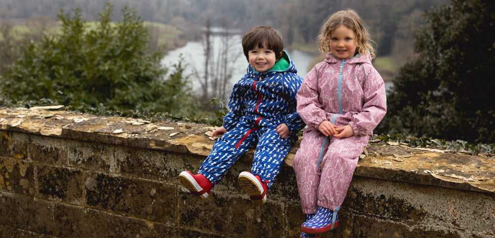 children sitting on brick wall with view of lake behind, wearing muddy puddles blue lightning puddlesuit and pink forest puddlesuit