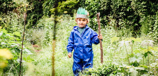 Child in blue waterproof suit exploring lush green outdoor setting, holding a stick