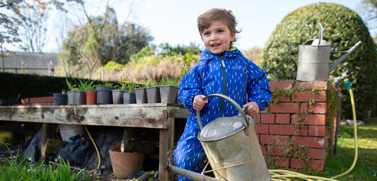 boy standing in garden holding large metal watering can, wearing vibrant blue muddy puddles puddlesuit