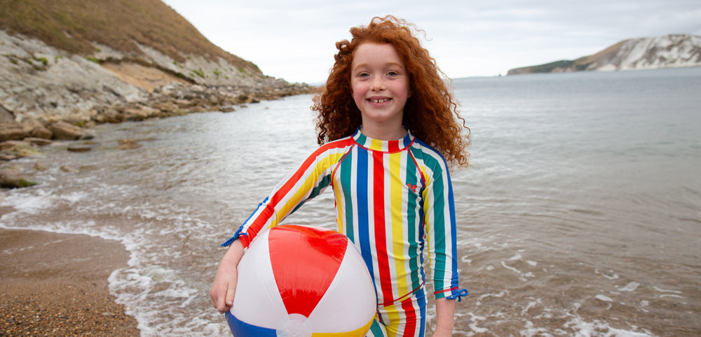 girl standing on a sandy beach, holding a beach ball, wearing striped sunsafe swimwear by muddy puddles
