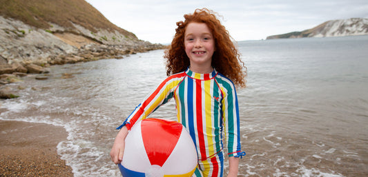 girl standing on a sandy beach, holding a beach ball, wearing striped sunsafe swimwear by muddy puddles