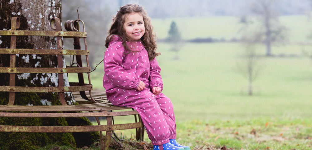 Smiling child in a pink waterproof suit sitting on a bench outdoors in a grassy field