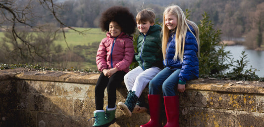 Three children in colorful waterproof jackets and boots sit on a stone wall outdoors