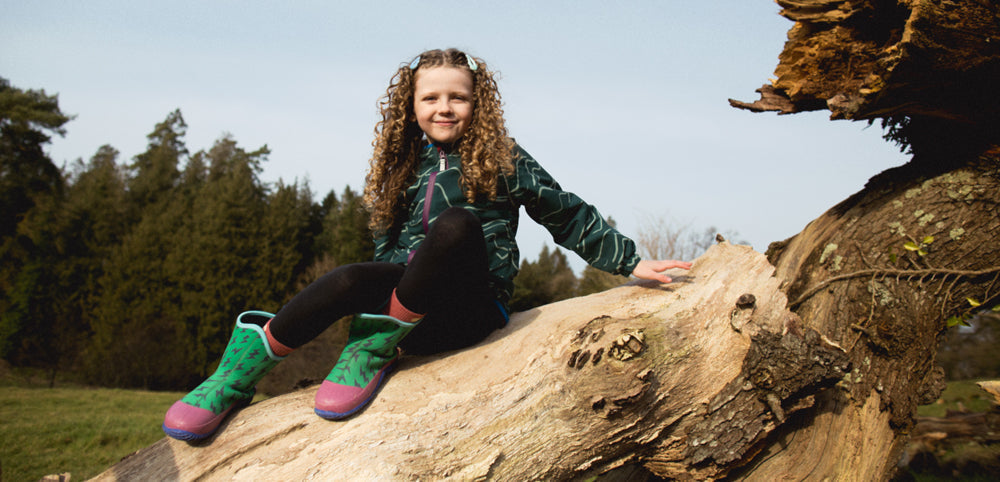 Child in waterproof jacket and boots sitting on fallen tree outdoors