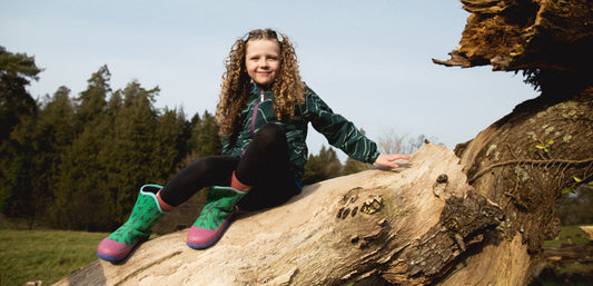 Child in waterproof jacket and boots sitting on fallen tree outdoors