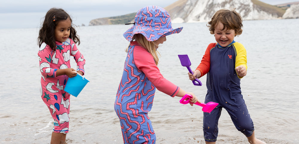 children playing in the sea with buckets and spades, wearing colourful muddy puddles surfsuits and sun hats