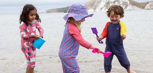 children playing in the sea with buckets and spades, wearing colourful muddy puddles surfsuits and sun hats