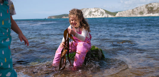 girl sitting on a rock in the sea holding seaweed, wearing muddy puddles pink waterproof dungarees and a white tshirt