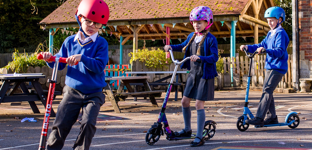 Children in school uniforms and helmets riding scooters outdoors on playground