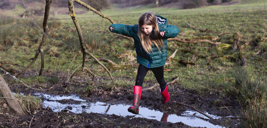girl jumping across muddy puddle wearing red wellies and green waterproof puffer jacket