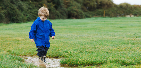 boy standing in puddle wearing muddy puddles blue waterproof jacket and navy waterproof trousers