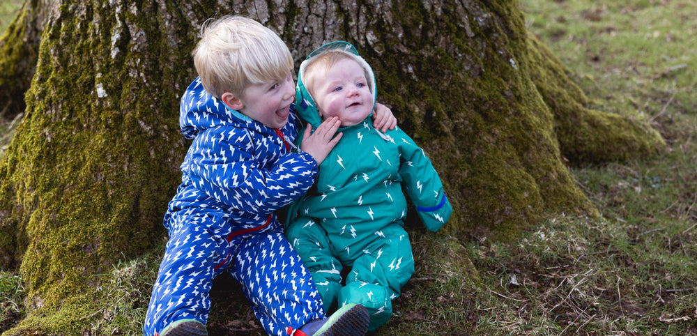 toddler and baby sat at bottom of tree wearing muddy puddles waterproof puddle suits
