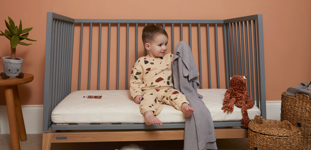 toddler sitting in grey cot on naturalmat mattress holding grey blanket