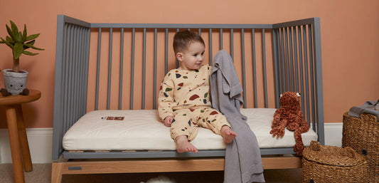 toddler sitting in grey cot on naturalmat mattress holding grey blanket