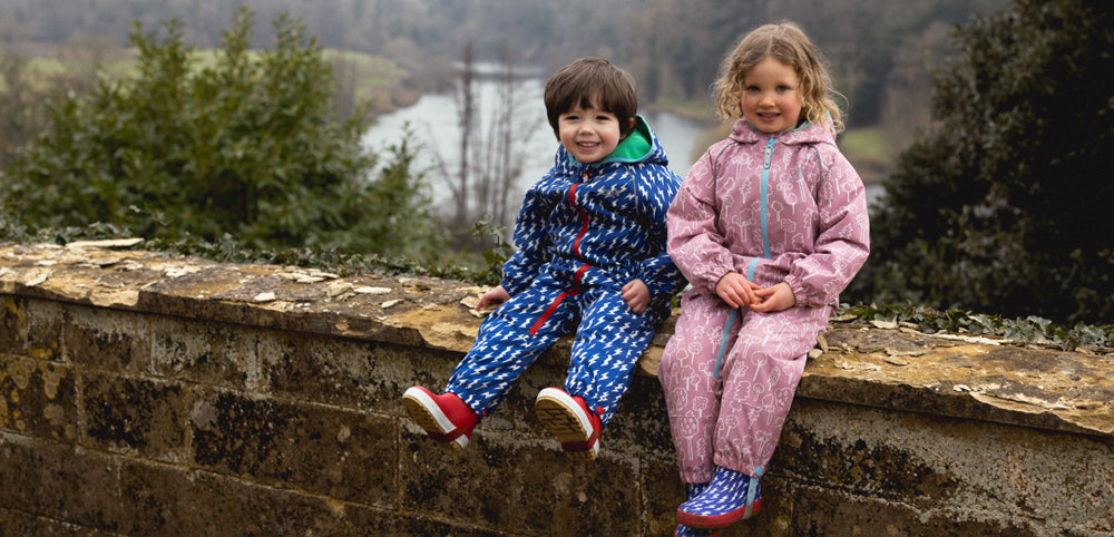 Two children in colorful waterproofs sitting on a stone wall outdoors near greenery