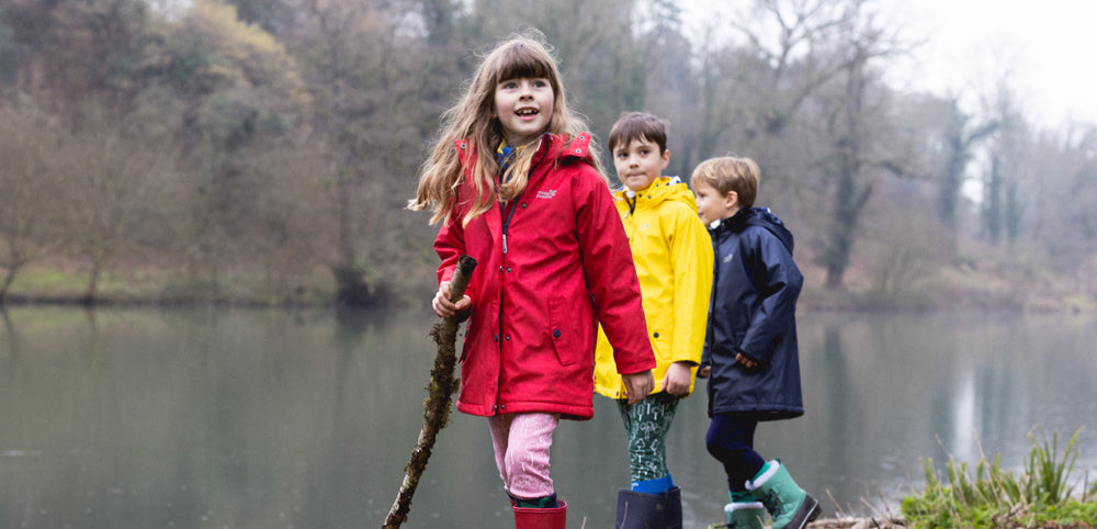 children stood by lake wearing red yellow and navy waterproof jackets, holding a large stick with trees in background
