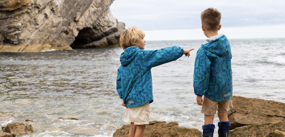two boys on rocky beach pointing out to sea, wearing vibrant green muddy puddles raincoats