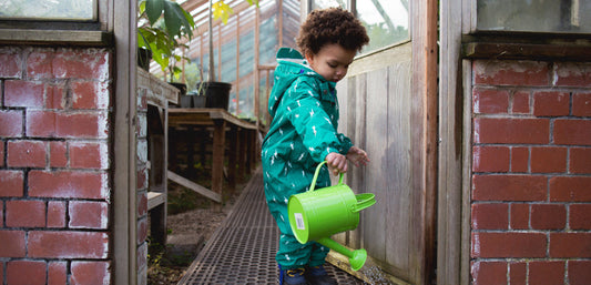 Child in green waterproof suit watering plants in a greenhouse