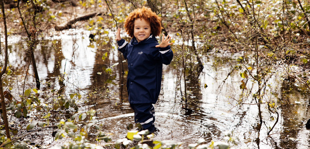 Child in navy waterproof rain suit playing outdoors in woodland puddle