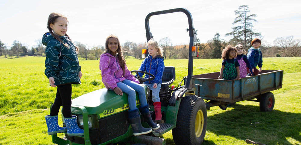 children sitting on green tractor and trailer, wearing colourful muddy puddles waterproofs
