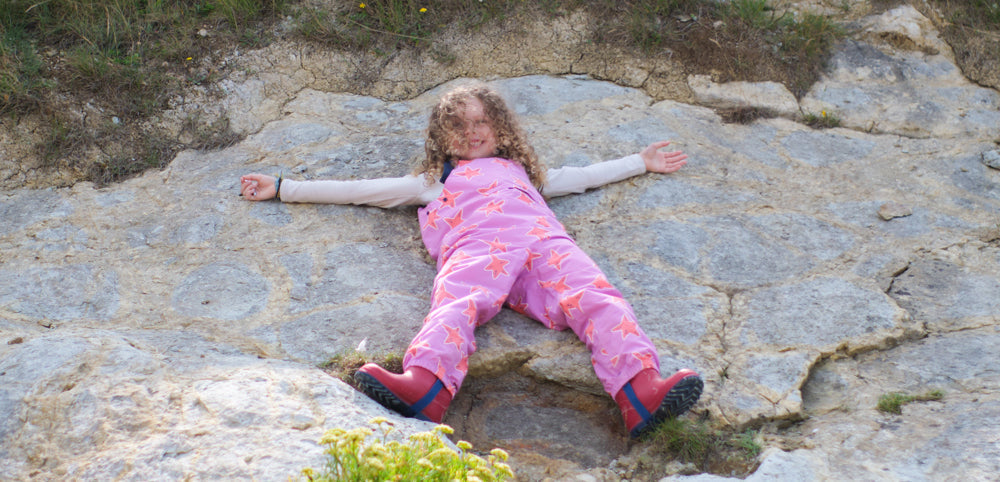 girl lying down on rocky surface wearing muddy puddles pink waterproof dungarees and red wellies