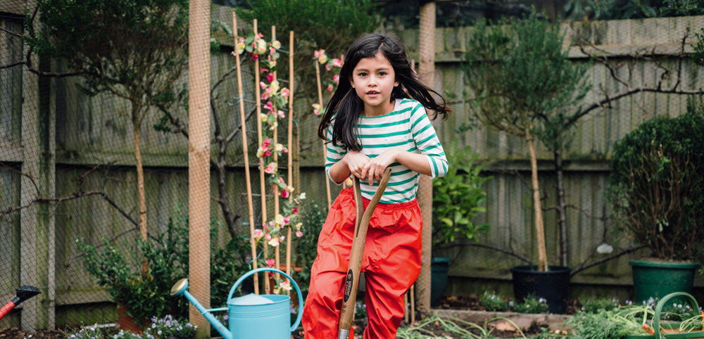 Young girl in a home garden with a garden spade, wearing durable, bright clothing suitable for outdoor play, surrounded by potted plants and a watering can.