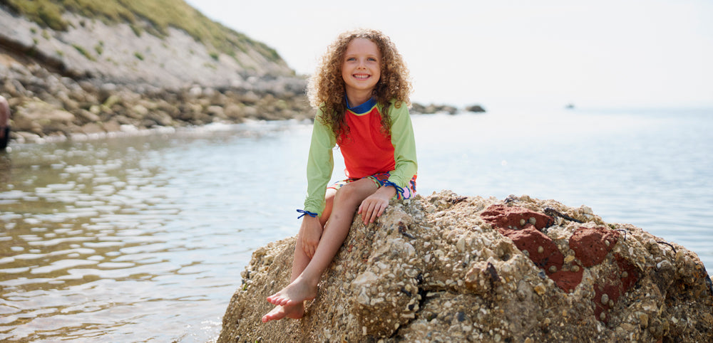 Smiling child in colorful waterproofs sitting on a rock by the sea on a sunny day
