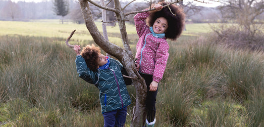 Children in waterproof jackets climbing a tree outdoors in grassy countryside.