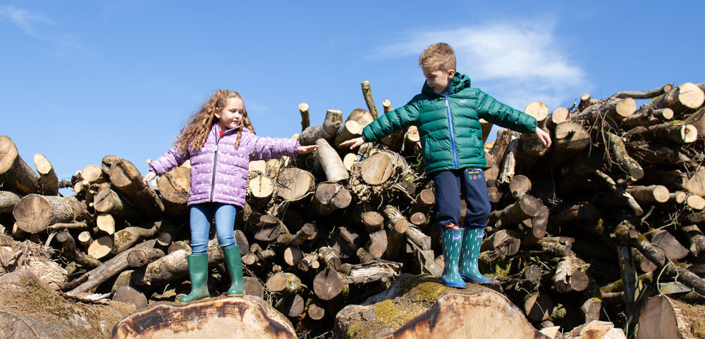 boy and girl standing on tree logs, reaching for each others hand, wearing muddy puddles green and lilac puffers