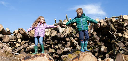 boy and girl standing on tree logs, reaching for each others hand, wearing muddy puddles green and lilac puffers