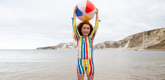 girl stood in the sea holding beach ball over her head, wearing striped muddy puddles swimwear