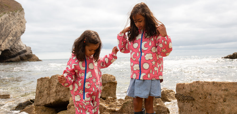 girls standing in rock pools by the sea, looking down at their feet, wearing vibrant pink muddy puddles waterproofs