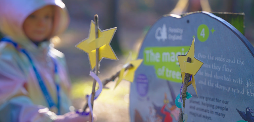 Child in hooded waterproof jacket holding star wands outdoors near forest activity sign