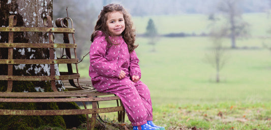 girl sat on bench under tree wearing pink muddy puddles scampsuit