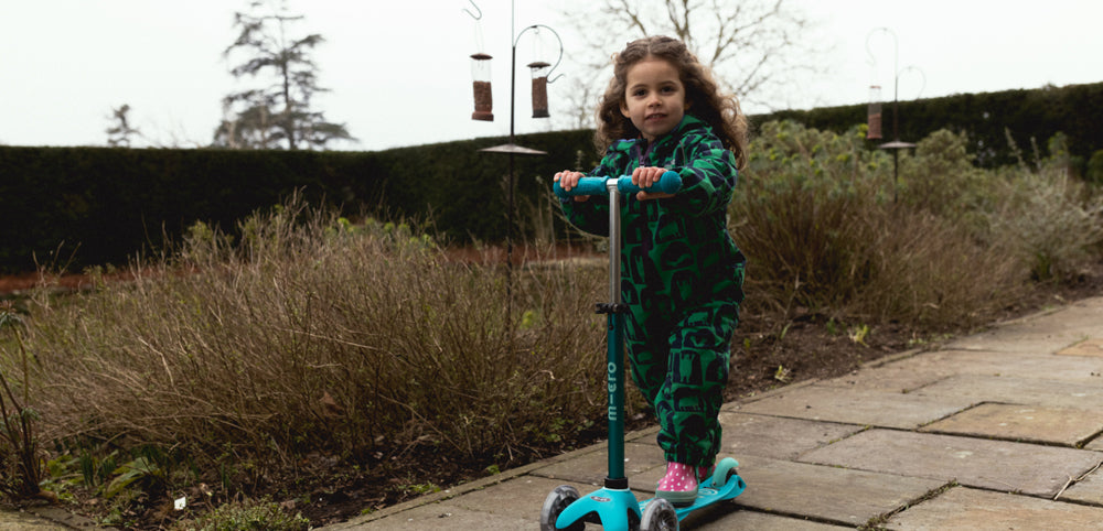 Child in green waterproof suit riding blue scooter outdoors on a garden path